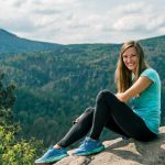 Smiling woman in blue sneakers sitting on a rock with mountain landscape in Jonsdorf, Germany.